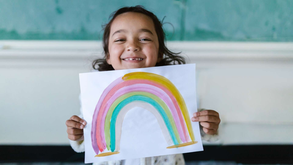 Smiling child holding a rainbow painting during a classroom art activity.