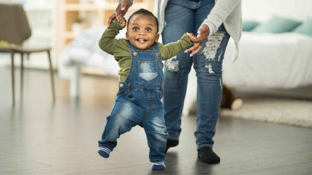 Smiling young child practicing walking while holding a caregiver’s hands for support.