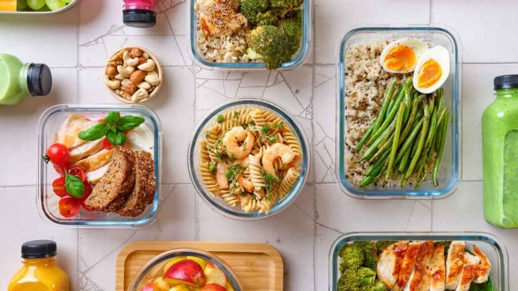 Healthy prepared meals with vegetables, fruit, pasta, rice, eggs, and drinks arranged in containers.